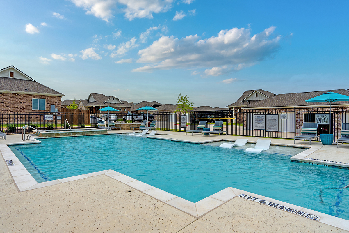 Outdoor swimming pool with lounge chairs and umbrellas, surrounded by a fence. Suburban-style houses are visible in the background under a partly cloudy sky. 3 FT 6 IN NO DIVING is marked on the pool edge.
