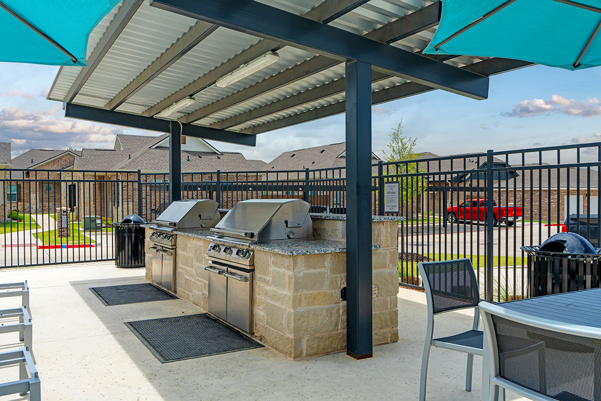 Outdoor kitchen with three stainless steel grills under a metal canopy, surrounded by stone counters, tables, chairs, and umbrellas. A fenced area with parked cars and houses is visible in the background.