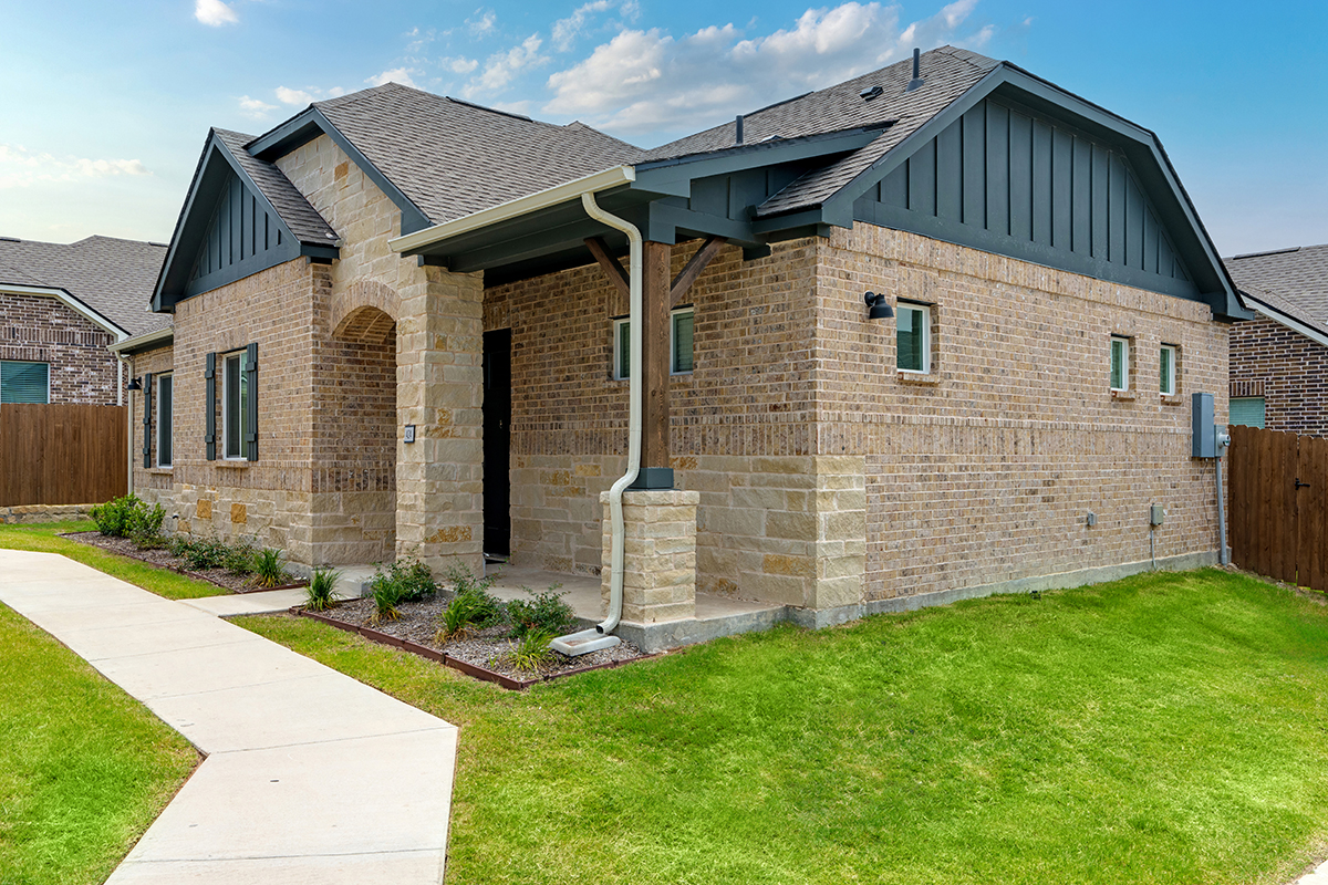A modern, single-story brick house with gray trim, a covered entryway, and well-maintained lawn under a blue sky. A concrete sidewalk curves along the front and side of the home.