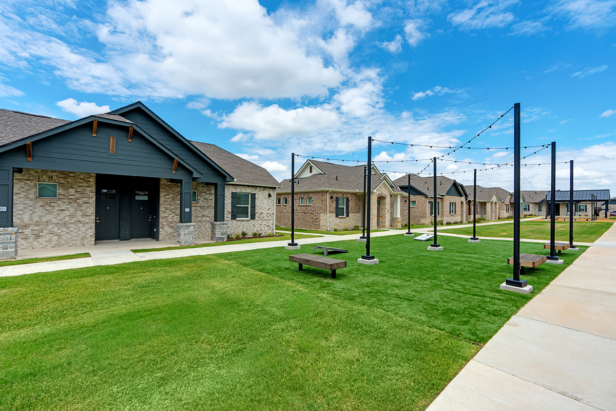 Row of modern brick houses with a grassy area, benches, and string lights on poles, under a partly cloudy blue sky. A sidewalk runs alongside the homes.