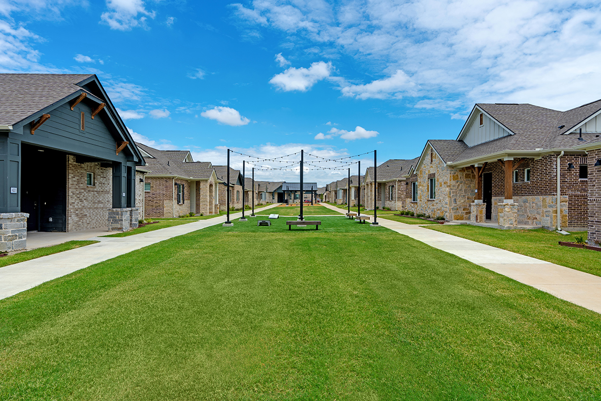 A grassy courtyard between rows of single-story brick and stone houses, with walkways on each side and string lights hanging above a central volleyball net. Blue sky with scattered clouds is visible.