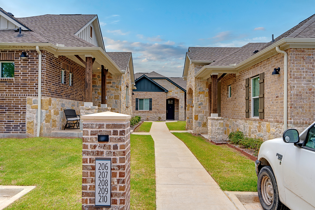 A sidewalk runs between brick buildings with stone accents. A white pickup truck is parked on the right, and a brick marker shows unit numbers 206–210. The sky is blue with scattered clouds.