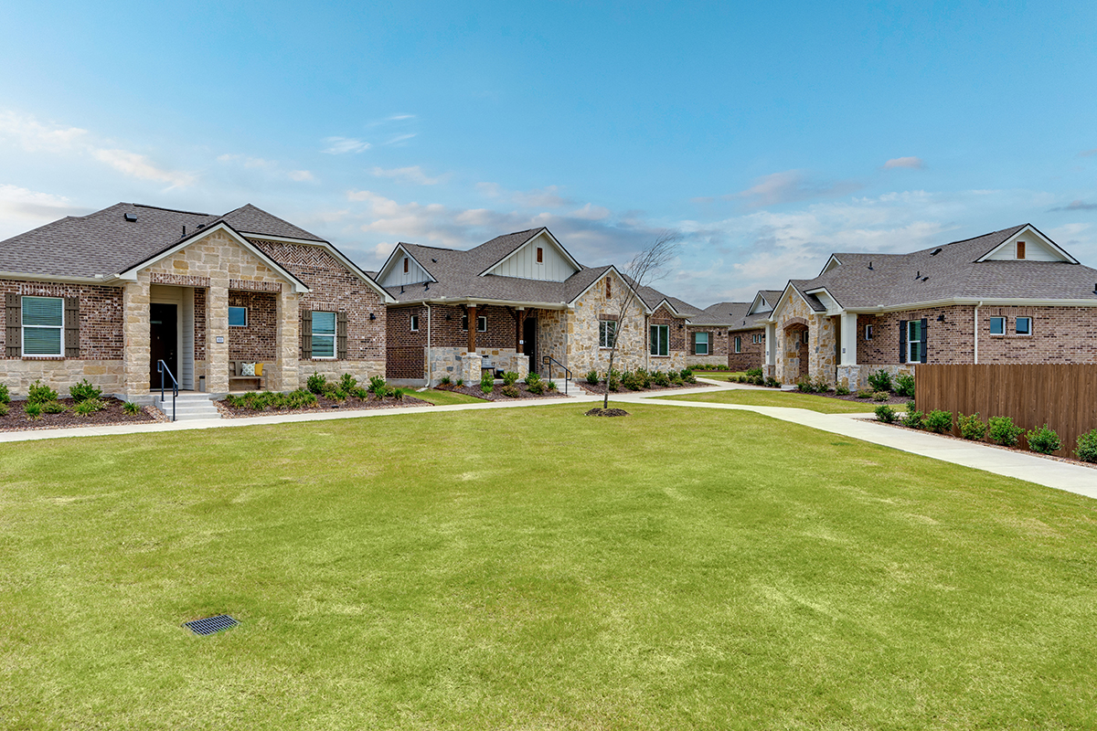 A row of modern, single-story brick houses sits along a wide, neatly trimmed green lawn under a blue sky with light clouds.