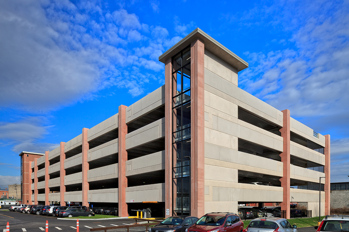 A multi-level parking garage with red vertical beams and beige concrete walls sits under a blue sky with scattered clouds; several cars are parked outside in the foreground.