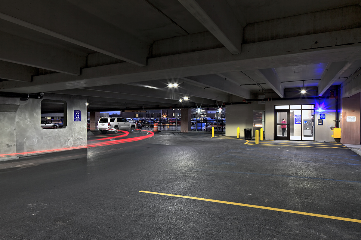 A dimly lit parking garage shows cars parked, a vehicle entering, and a booth with glass windows and a person inside near the entrance. Yellow lines and blue handicap spaces are visible on the asphalt.