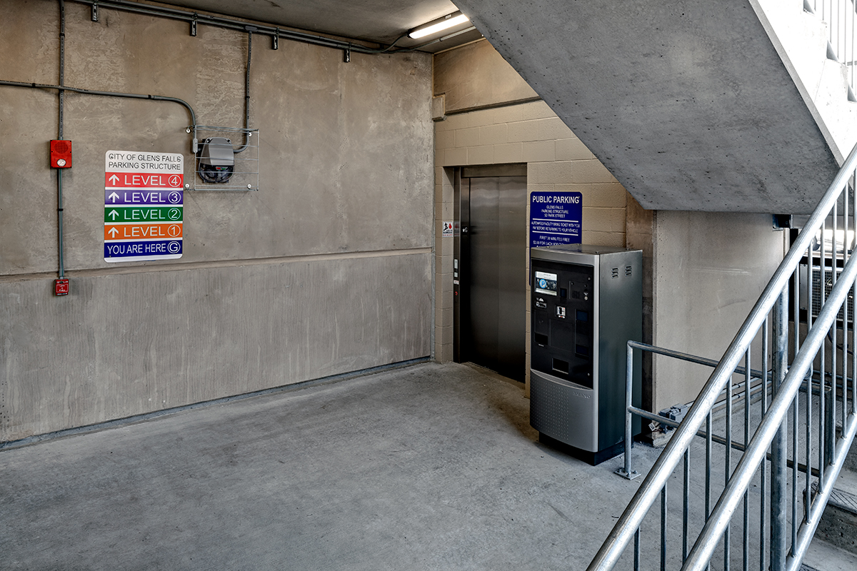 A parking garage corner with a payment machine, elevator, stairs, and colorful signs showing parking levels and locations. Walls and floor are concrete.
