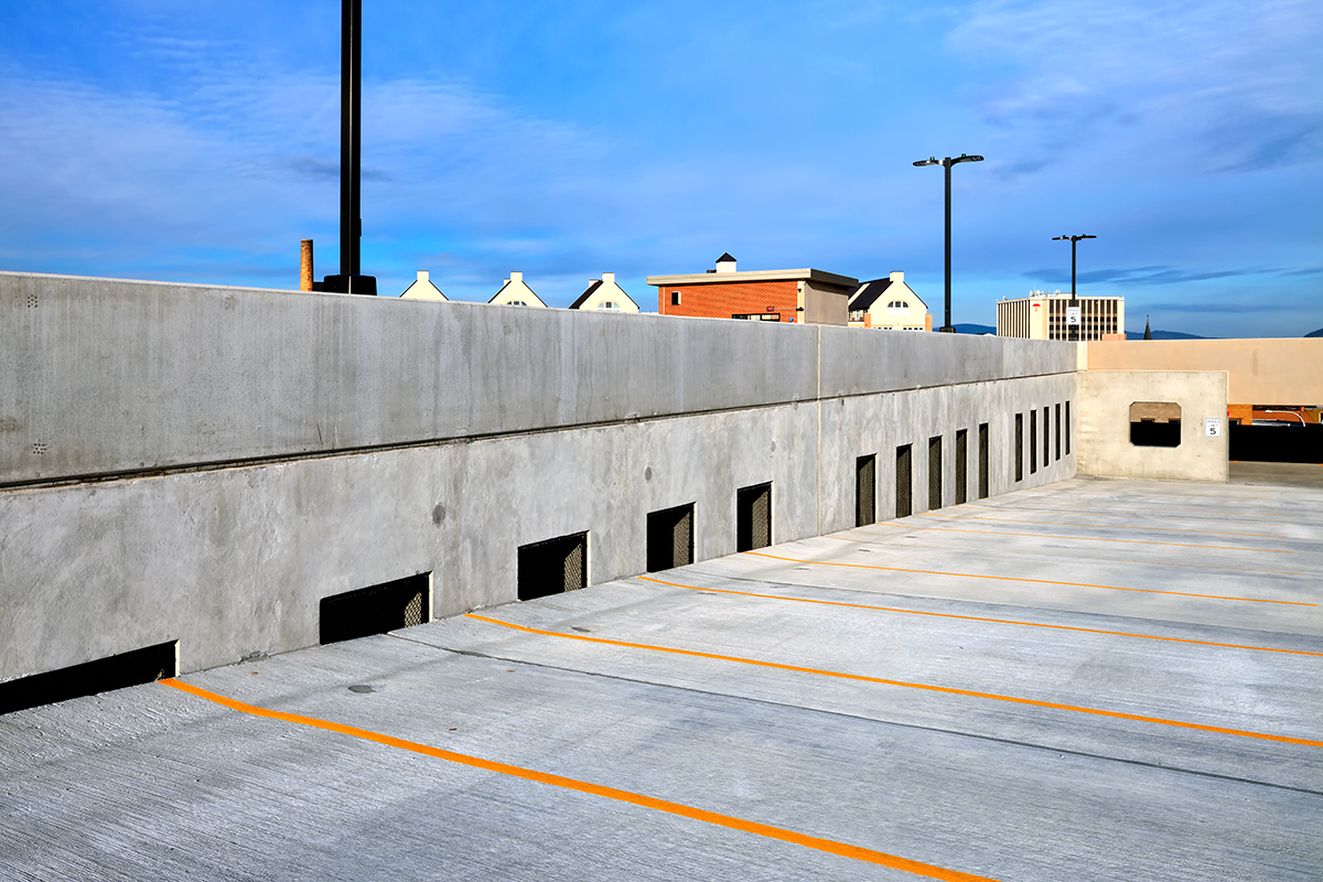 Empty rooftop parking lot with angled spaces, concrete barriers, and yellow lines. Buildings with white roofs are visible in the background under a bright blue sky.