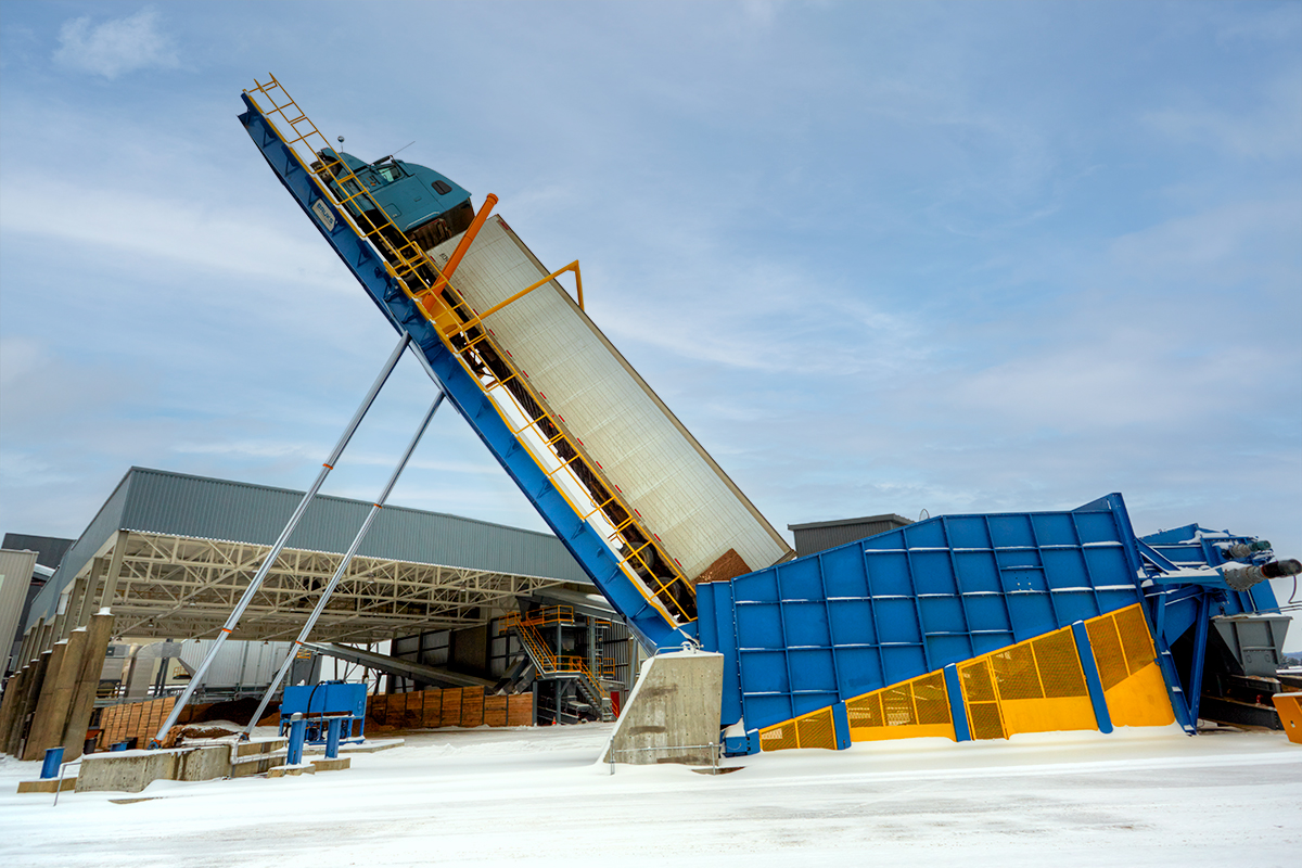 A blue industrial dump truck is tilted upward on a large, blue and yellow metal platform near an industrial facility on a snowy day.