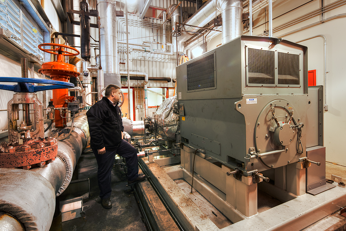 A person in a black jacket inspects large industrial equipment and pipes inside a brightly lit facility with metal structures and valves.