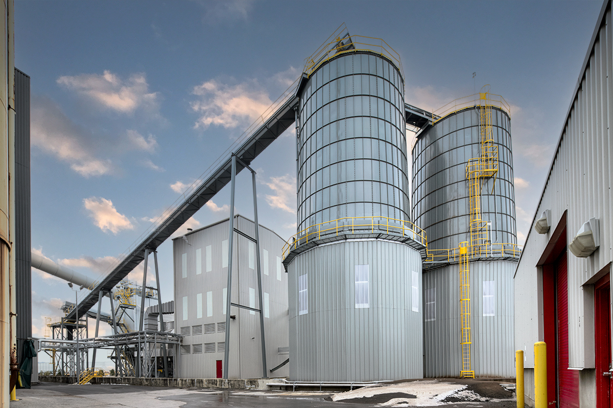 Large industrial silos with metal exteriors and yellow railings are connected by conveyors next to a factory building under a partly cloudy sky. Snow and patches of wet pavement are visible at the base.