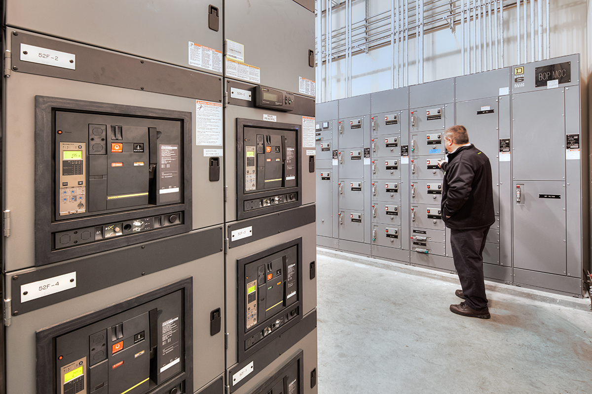 A man in a black jacket stands in a control room, operating a panel on a wall lined with electrical control cabinets and circuit breakers. The room has gray floors and overhead wiring.
