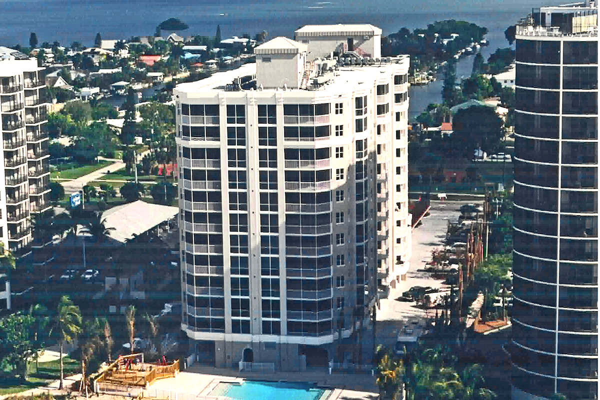A tall white apartment building with balconies stands near a swimming pool. Other high-rise buildings, palm trees, and water are visible in the background under a clear sky.