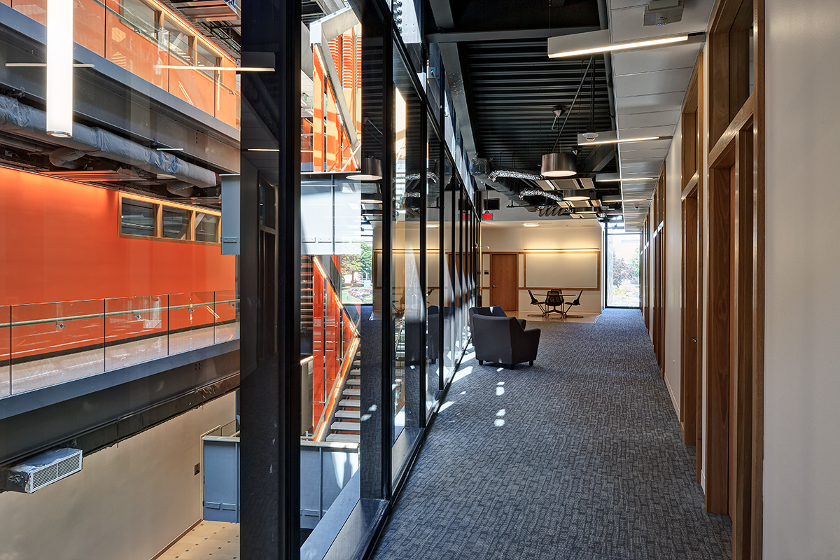 Office hallway with glass walls overlooks an open atrium with orange accents and exposed stairs. Two chairs and a table sit by windows at the end of the corridor. Ceiling features exposed pipes and modern lighting.