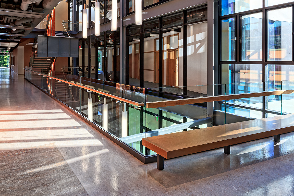 A modern building interior with large windows, exposed ceilings, glass railings, wooden benches, and natural light streaming in, creating an open and airy atmosphere.