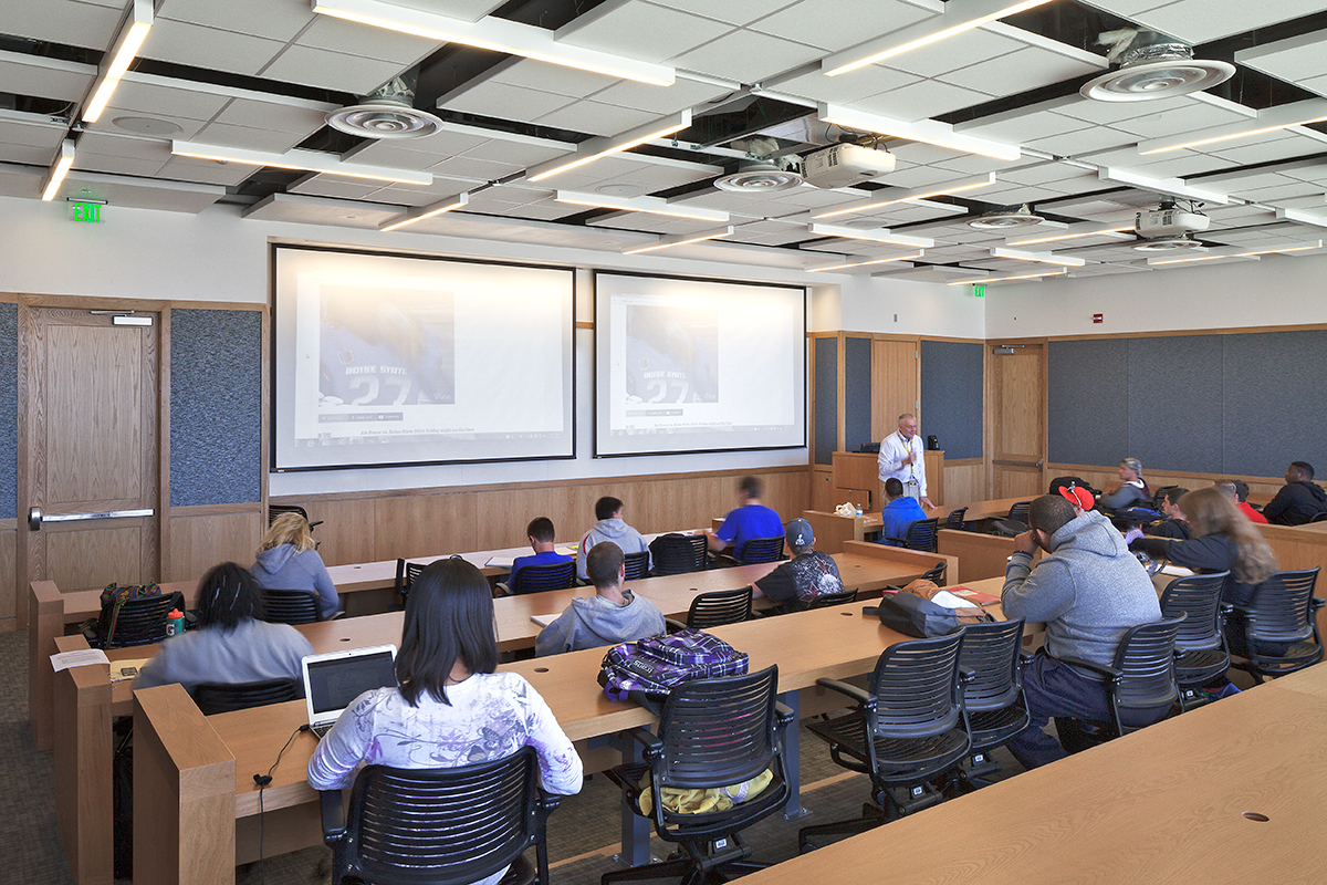 A classroom with students seated at desks facing forward, watching a lecturer standing near two large projection screens displaying slides. The room has modern lighting and wood accents.