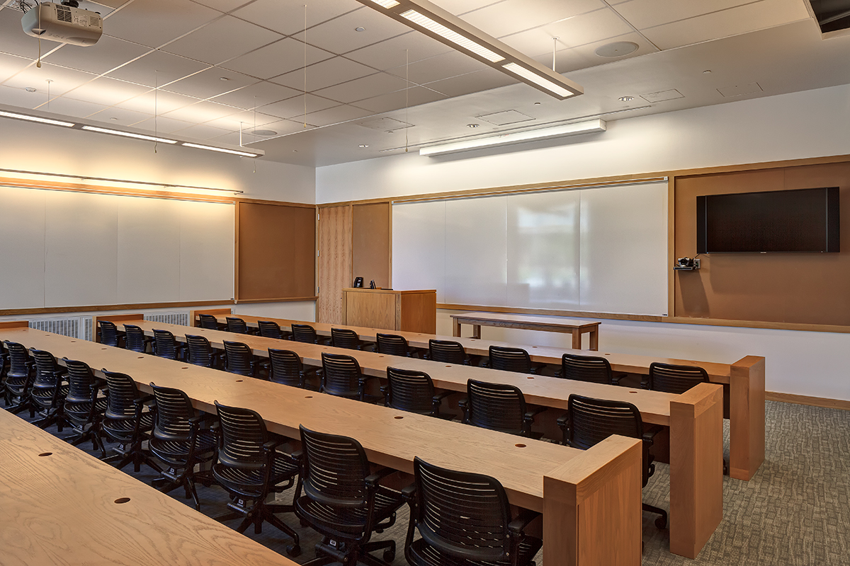 A modern, empty classroom with rows of wooden desks, black chairs, a whiteboard, a podium, a wall-mounted TV, and overhead lighting.