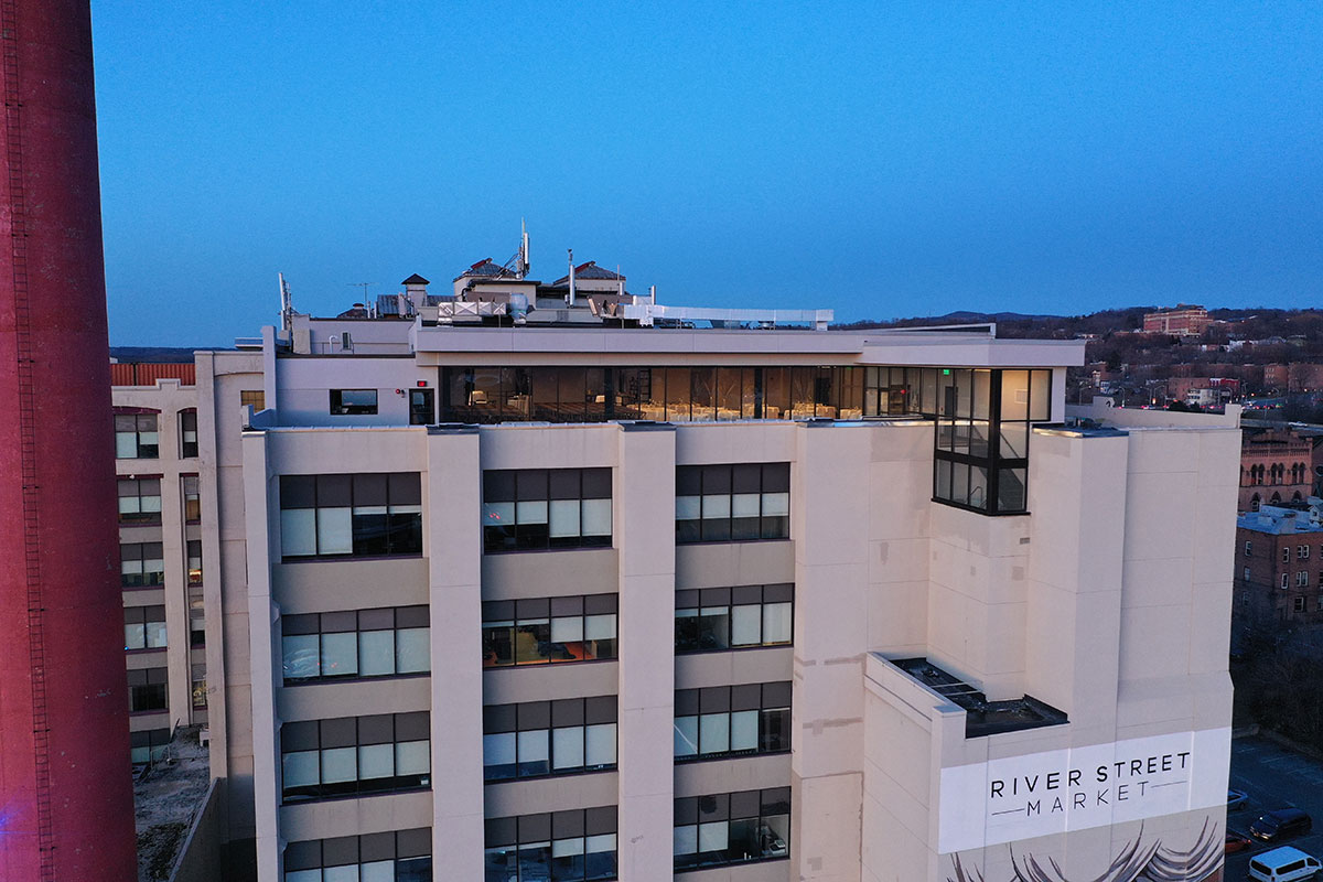 A modern multi-story building at dusk with large windows, rooftop seating, and a River Street Market sign on the exterior. City buildings and a red smokestack are visible in the background.