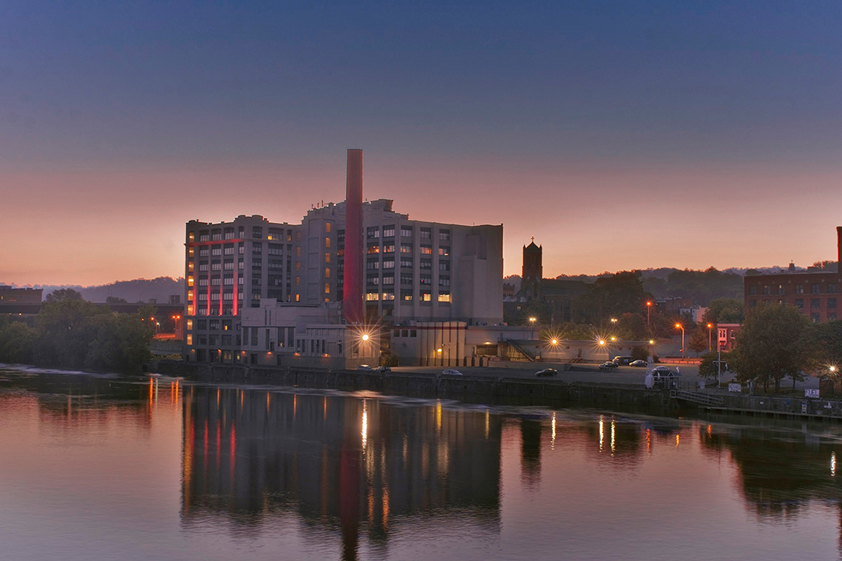 A modern building with a tall smokestack is reflected in a calm river at sunset, with city lights and trees visible along the waterfront.