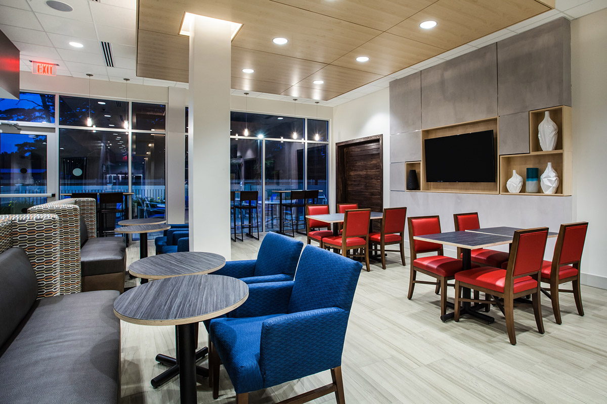 Modern hotel lounge with blue and red chairs, round and square tables, large windows, a wall-mounted TV, and decorative shelves with white vases.