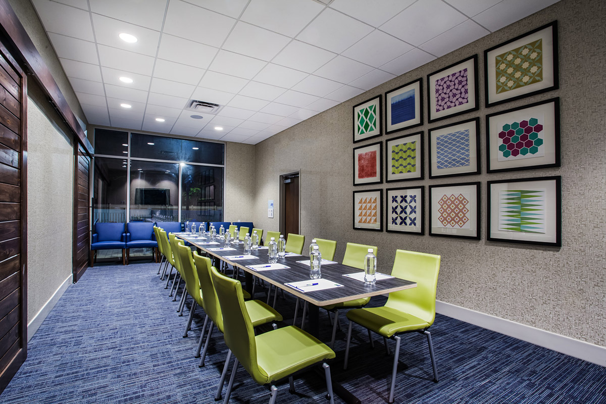 A modern conference room with a long table, green chairs, water bottles, notepads, and pens. Framed geometric art decorates the wall, and blue armchairs sit by large windows.