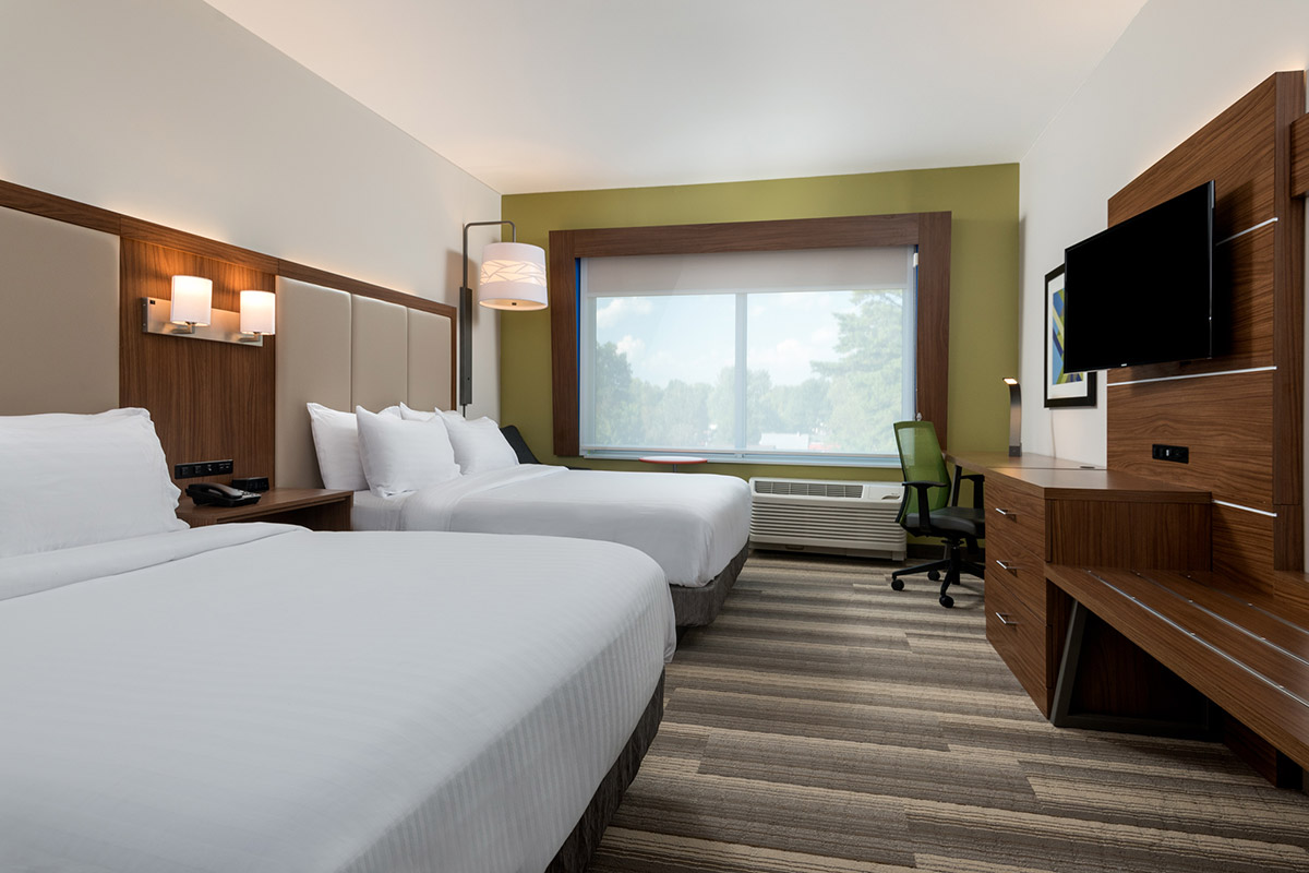 Modern hotel room with two queen beds, white linens, a wooden desk with a green swivel chair, flat-screen TV, large window with a view of trees, and soft lighting.