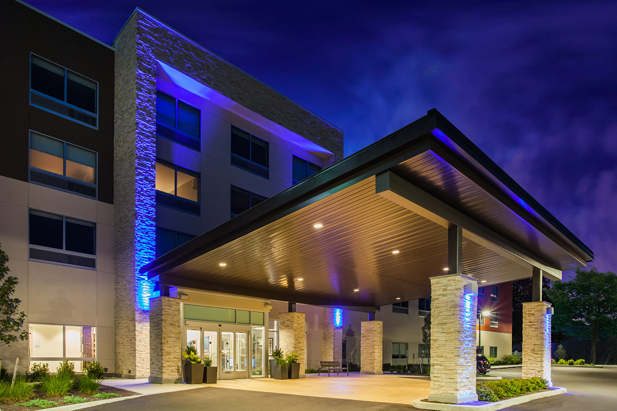 Modern hotel entrance at night, featuring a large covered driveway, tall glass doors, illuminated stone pillars, and blue accent lighting on the buildings exterior.