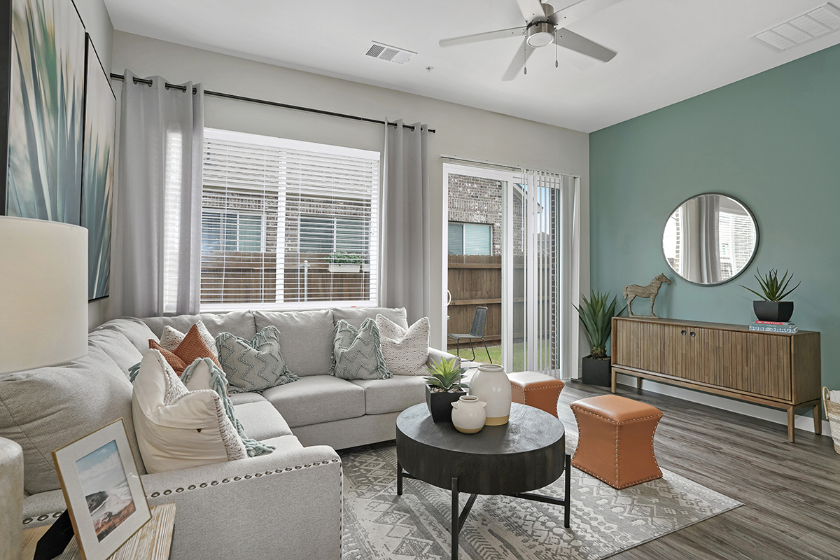 A modern living room with a gray sectional sofa, round black coffee table, light wood floors, green accent wall, round mirror, and plants. Large windows and sliding door bring in natural light.