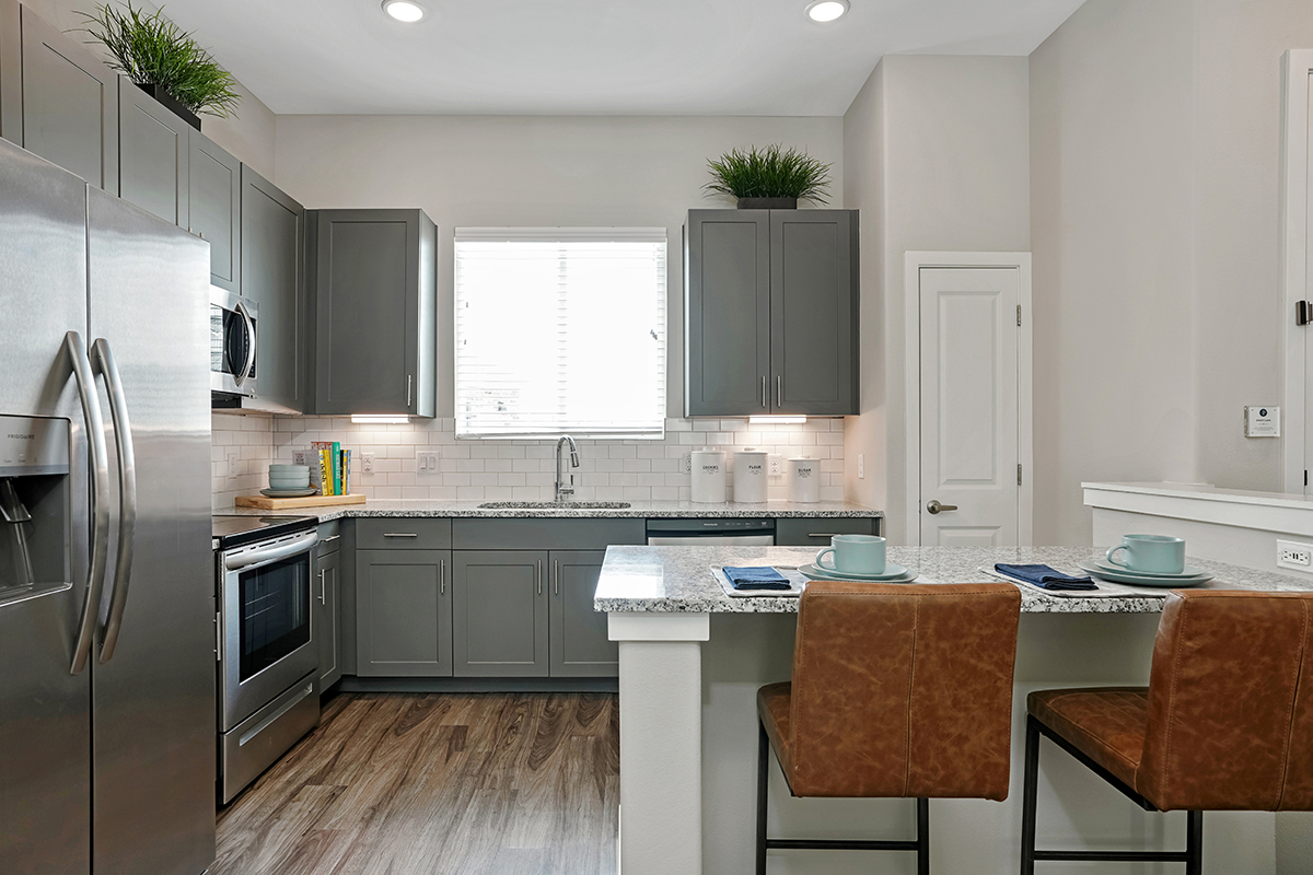 Modern kitchen with gray cabinets, stainless steel appliances, granite countertops, wooden floor, and two brown leather barstools set at a breakfast bar. Green plants sit on top of wall cabinets.