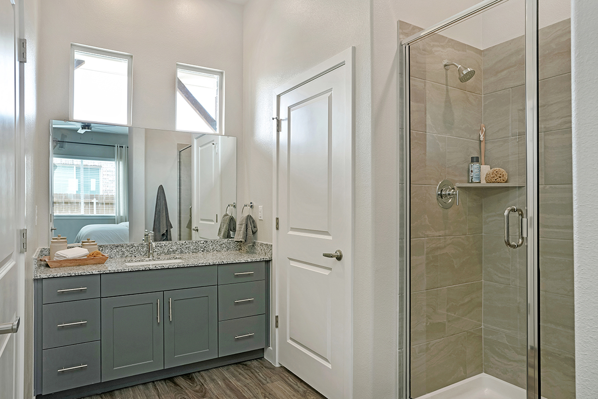 Modern bathroom with gray vanity, granite countertop, large mirror, glass shower with beige tiles, white walls, and wood-style flooring. Two small windows provide natural light. Towels and toiletries are neatly arranged.