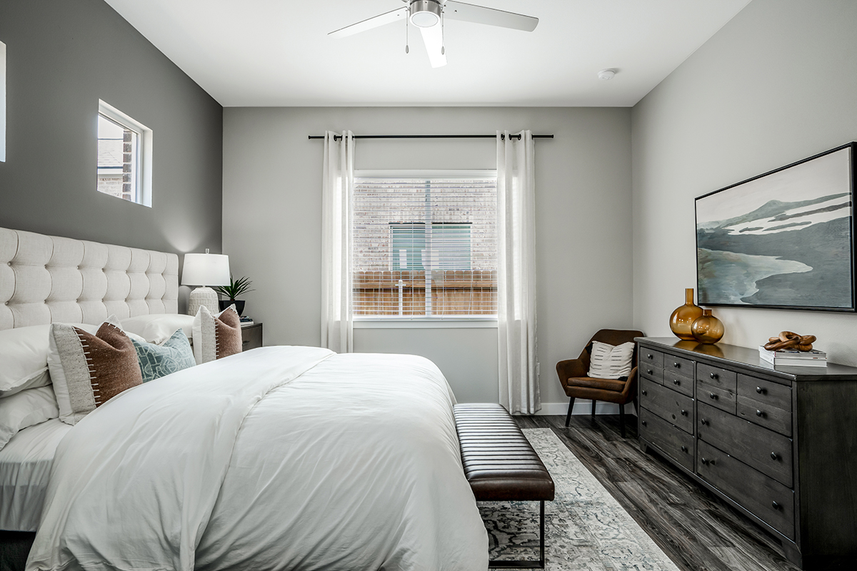 Modern bedroom with a large bed, bench, dresser, armchair, and decor. Sunlight streams through a window with white curtains, and a ceiling fan is above. Walls are light gray, and the floor is dark wood.