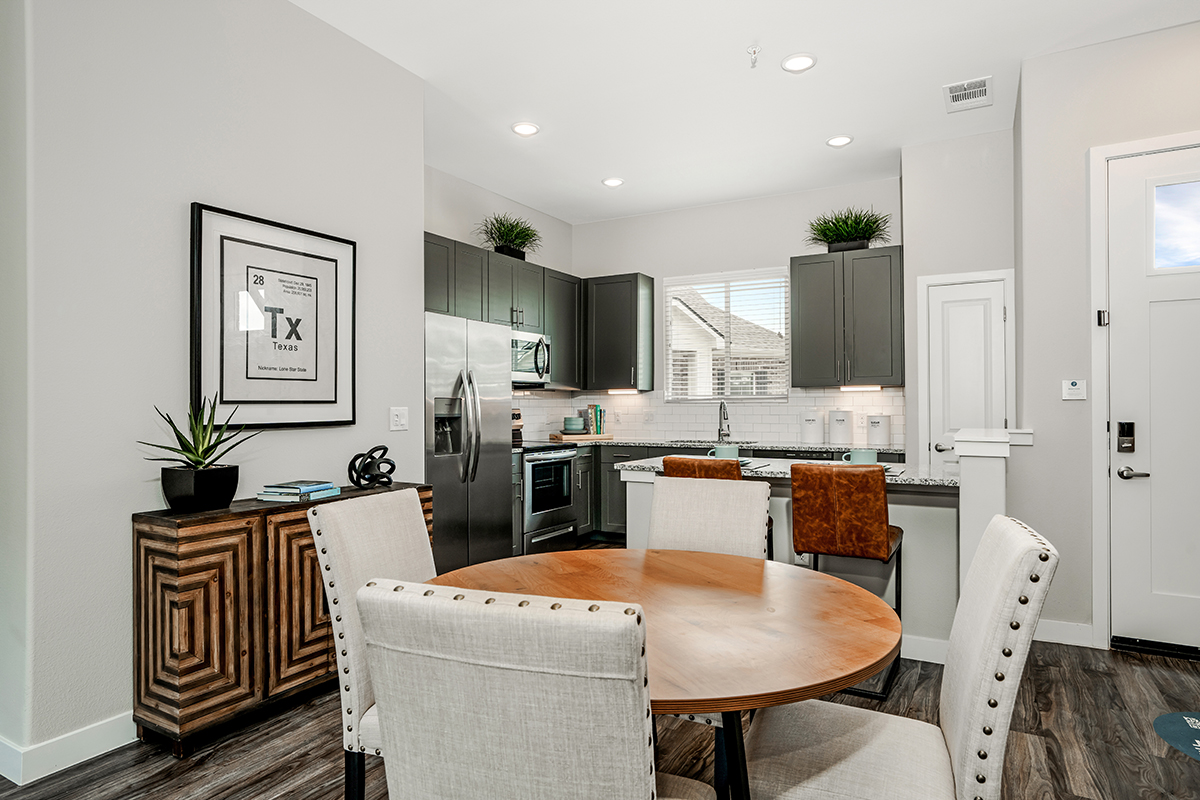 Modern kitchen and dining area with a round wooden table, upholstered chairs, dark cabinets, stainless steel appliances, and decorative plants on top of cabinets. Light walls, wood flooring, and wall art create a cozy atmosphere.
