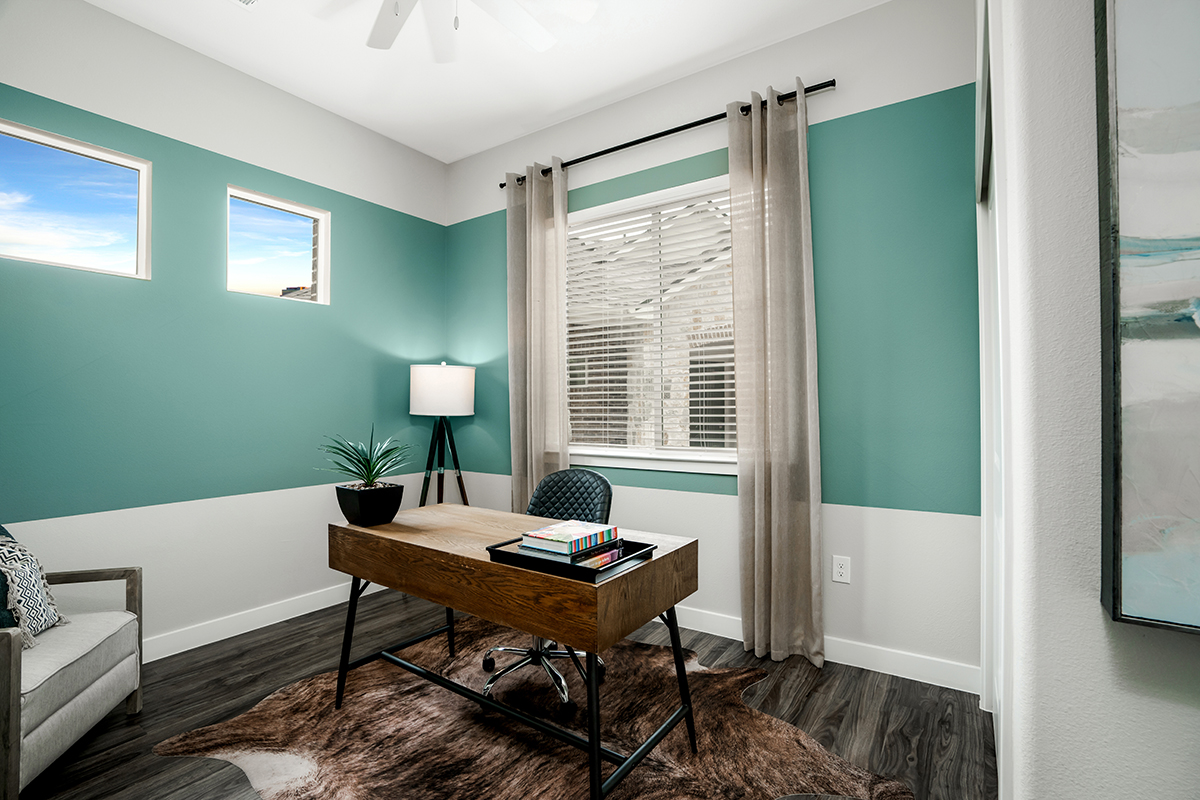 A modern home office with a wooden desk, black chair, lamp, plant, books, and large window with beige curtains. Walls are painted teal and white, and the floor has a dark wood finish with a brown rug.
