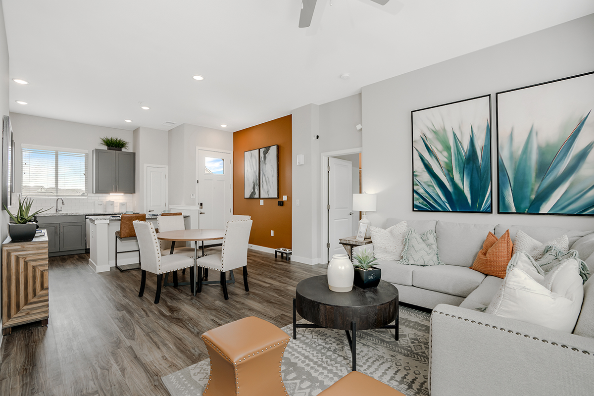 Modern open-concept living and dining area with gray sofa, orange accents, large plant artwork, and a kitchen with white cabinets, hardwood floors, and natural light from windows.