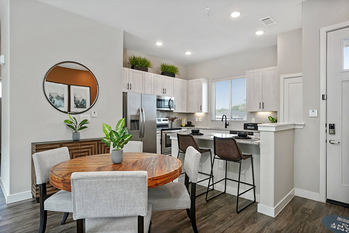 Modern kitchen and dining area with a round wooden table, four upholstered chairs, white cabinets, stainless steel appliances, barstools by a counter, green plants, and a round wall mirror reflecting framed art.