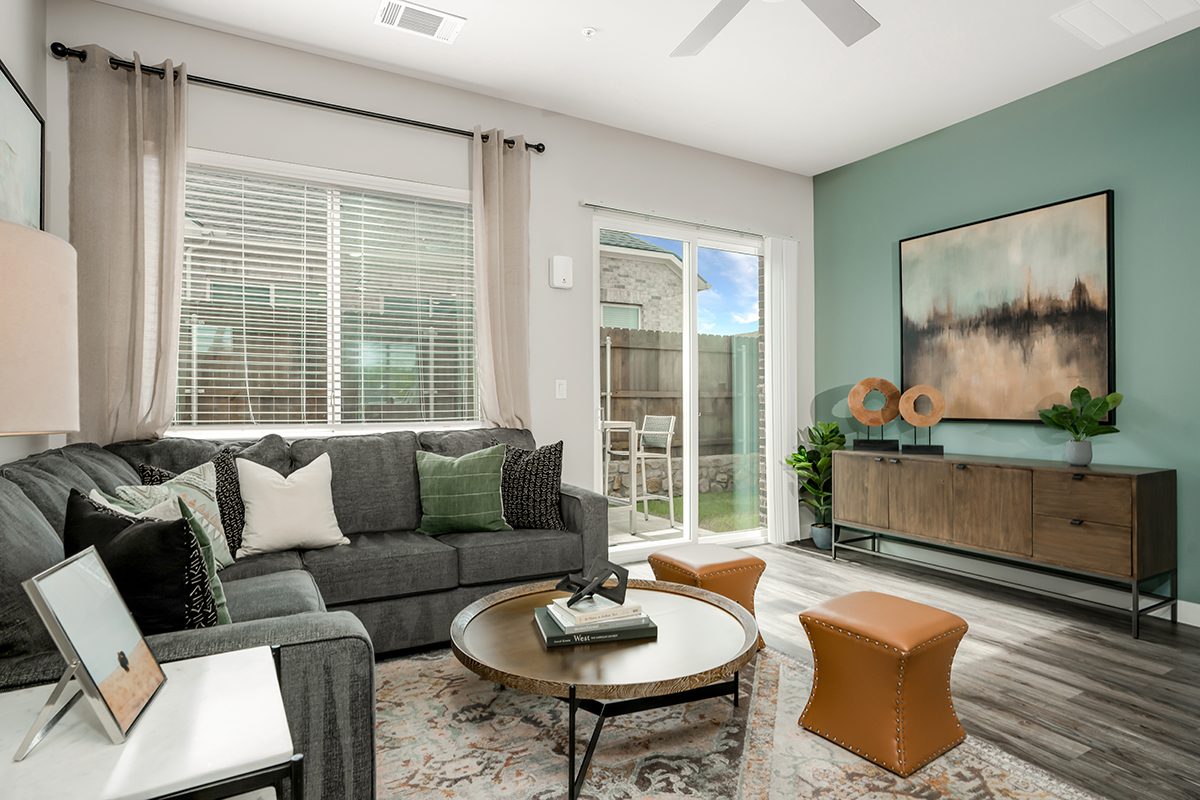 Modern living room with a gray sectional sofa, round coffee table, two brown ottomans, a wooden console, abstract wall art, large window, and sliding glass door leading to a patio. Decor includes plants and neutral-toned accessories.