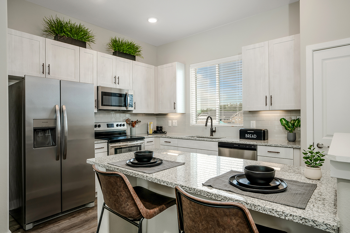 Modern kitchen with white cabinets, stainless steel appliances, granite countertops, two brown chairs, two black bowls on placemats, potted plants, and a window with blinds letting in natural light.