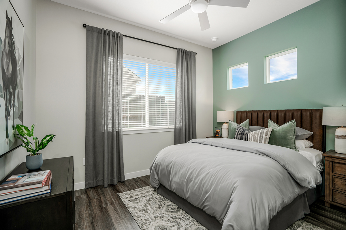 Modern bedroom with a green accent wall, two small windows, a large bed with gray bedding, two bedside tables with lamps, a plant on a dresser, and a window with gray curtains letting in daylight.