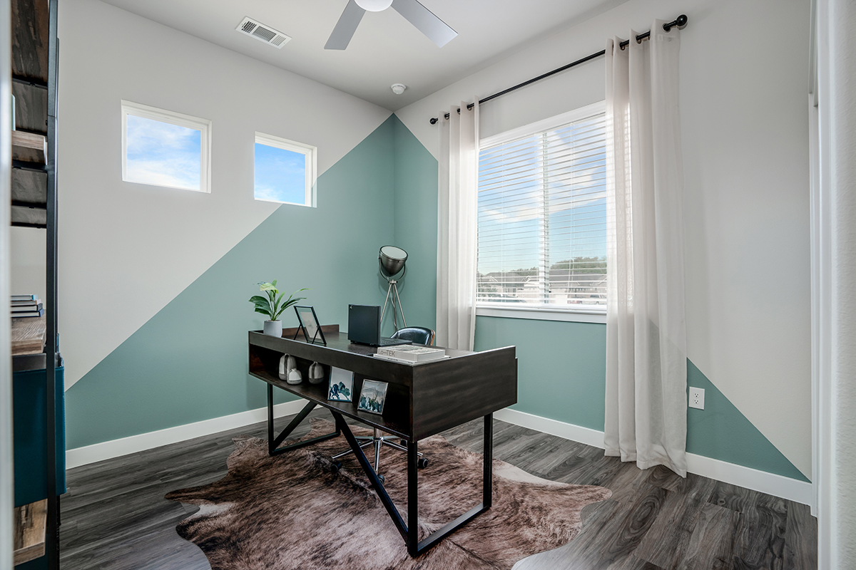 Modern home office with geometric light green and white walls, a dark desk, laptop, plant, lamp, and cowhide rug. Large window with blinds lets in natural light. Ceiling fan and hardwood floors complete the space.