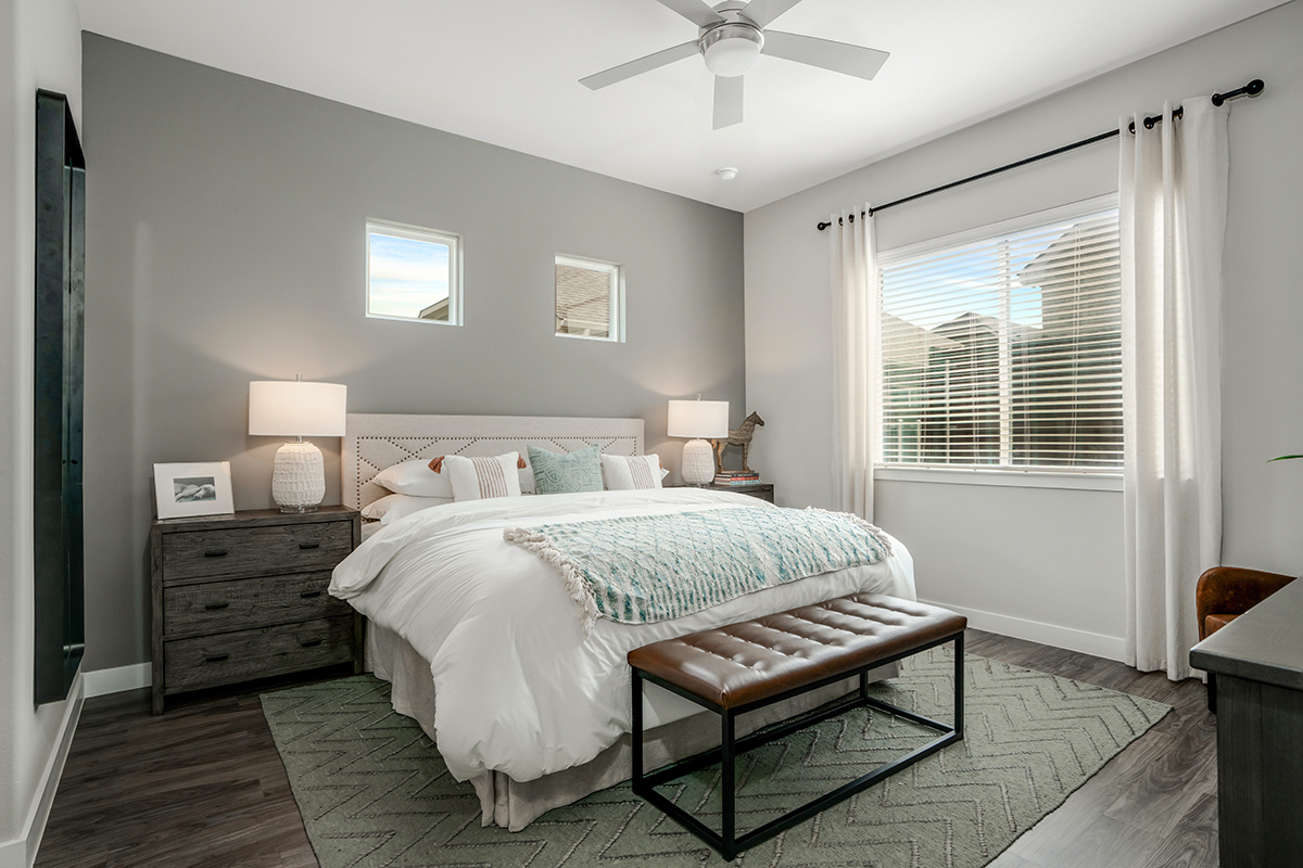 Modern bedroom with a gray accent wall, double nightstands, a large bed, two lamps, a bench at the foot of the bed, and a window with white curtains letting in natural light.