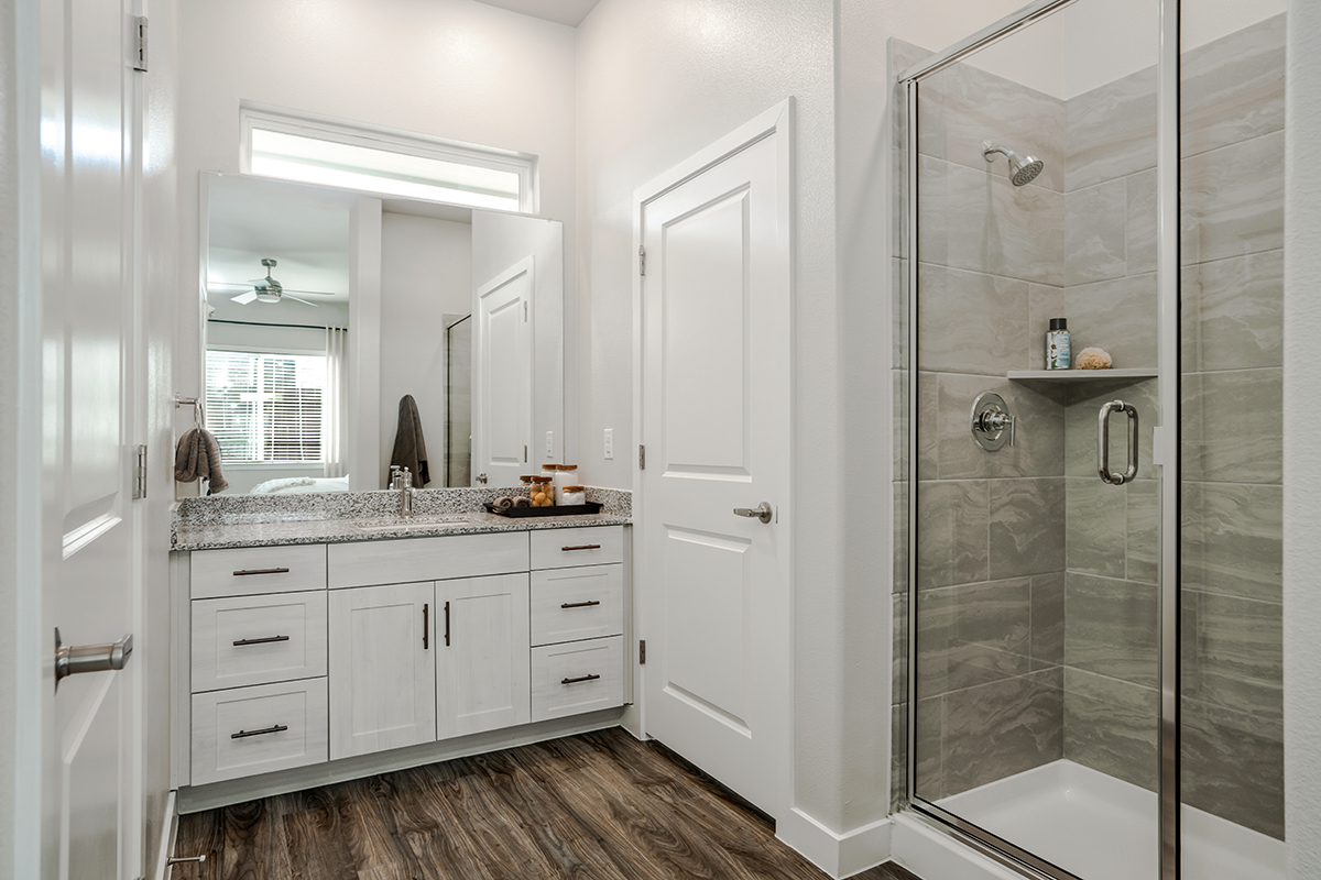 Modern bathroom with a glass shower, gray tile, white vanity with double sinks, granite countertop, and wood-look flooring. A mirror reflects a bedroom with a ceiling fan and large window.