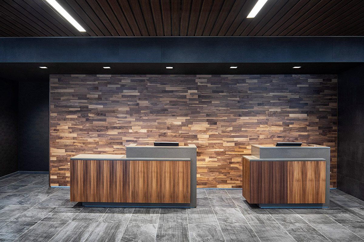 Two modern wooden reception desks sit in front of a decorative wood-paneled wall, with gray tile flooring and dark ceiling panels above, under bright rectangular lights.