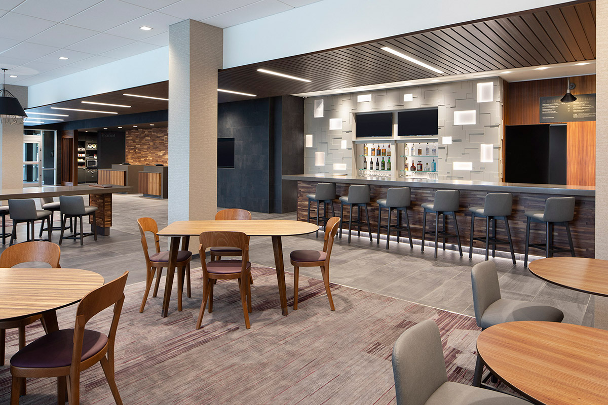 Modern hotel lounge with round wooden tables, cushioned chairs, and a bar area with high stools. The space features contemporary lighting, neutral tones, and a clean, open layout suitable for dining or socializing.