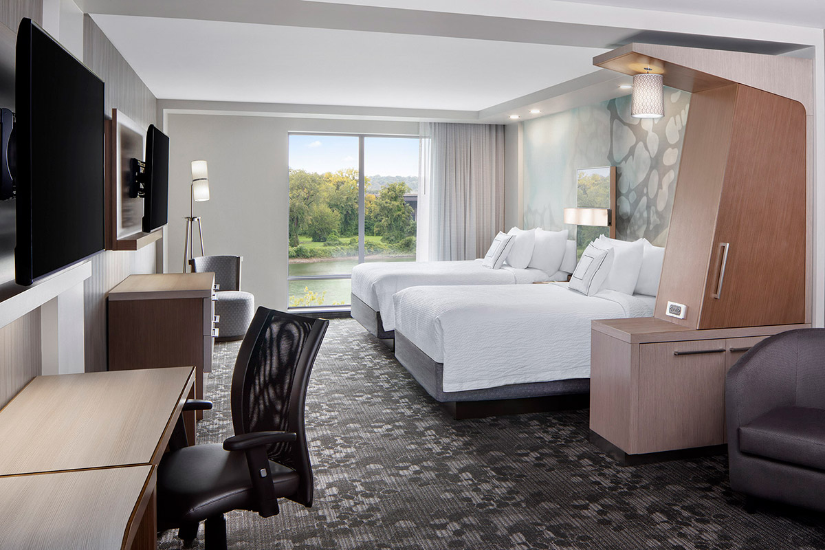 Modern hotel room with two beds, desk, office chair, TV, and armchair. Large window with a scenic view of trees and a river lets in natural light. Neutral colors and contemporary decor create a clean, inviting atmosphere.