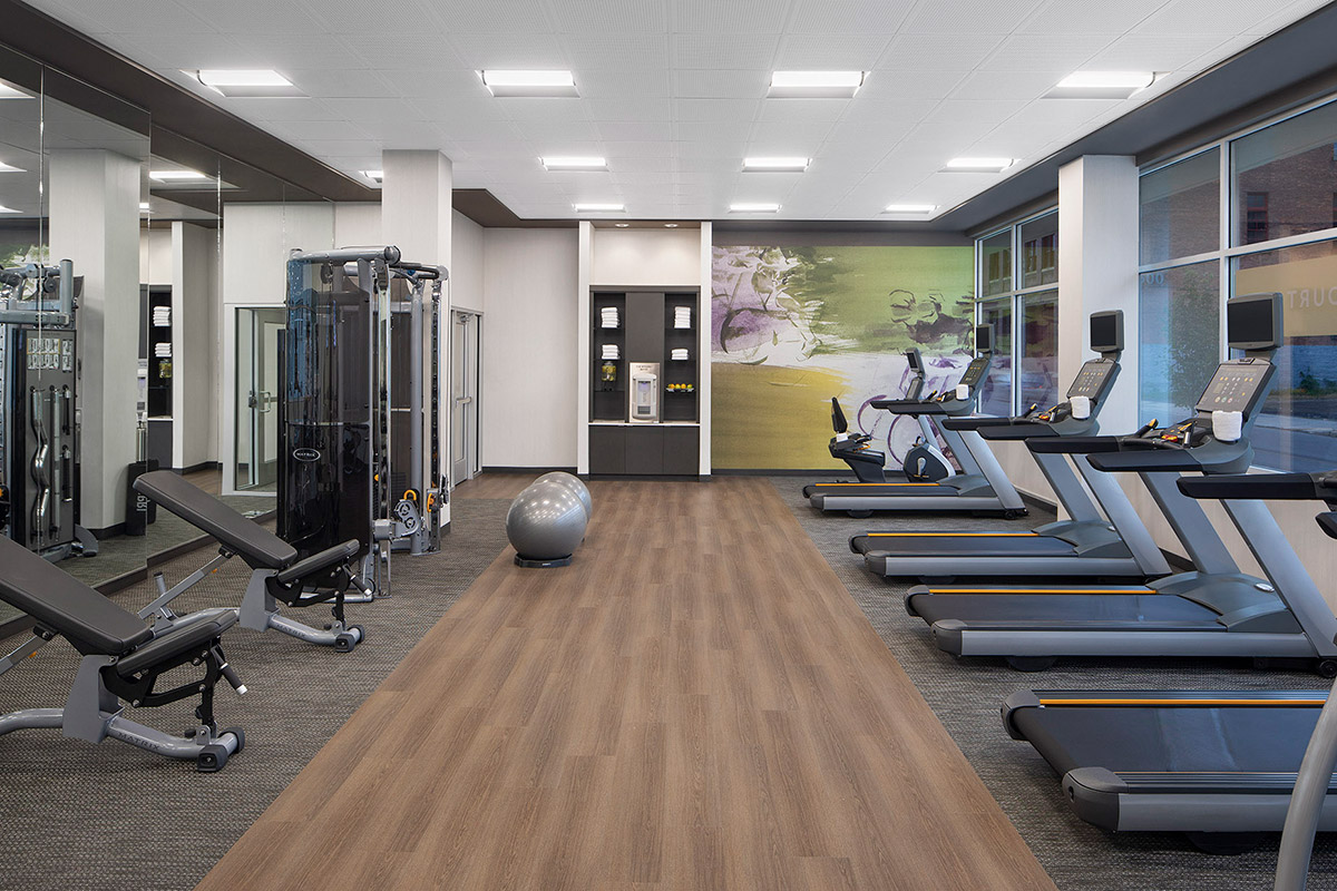 Modern gym with treadmills, weight benches, exercise machines, a stability ball, mirrors on one wall, and a mural on the far wall. The space has wood and carpet flooring with bright ceiling lights.