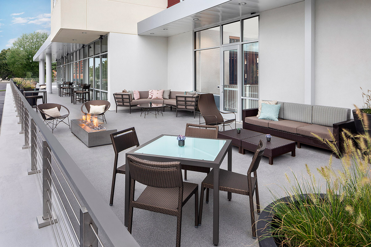 Modern outdoor patio with wicker chairs, sofas, tables, a fire pit, and decorative pillows. The area is adjacent to a building with large windows and surrounded by greenery.