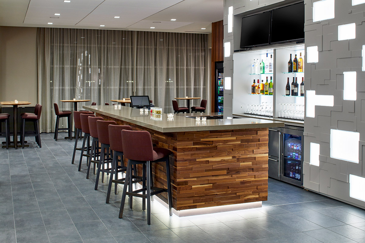 Modern bar area with a wooden counter, high chairs, shelves of liquor bottles, glassware, and soft lighting, surrounded by tables and chairs, in a clean, stylish setting.