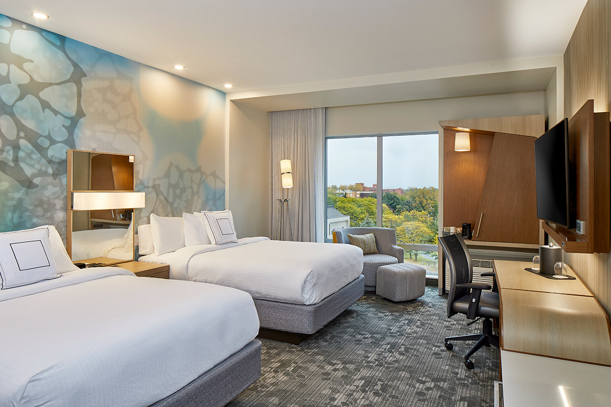 Modern hotel room with two double beds, a work desk with a chair, a wall-mounted TV, a lounge chair by a large window, and blue-patterned accent wall décor. Natural light fills the space.
