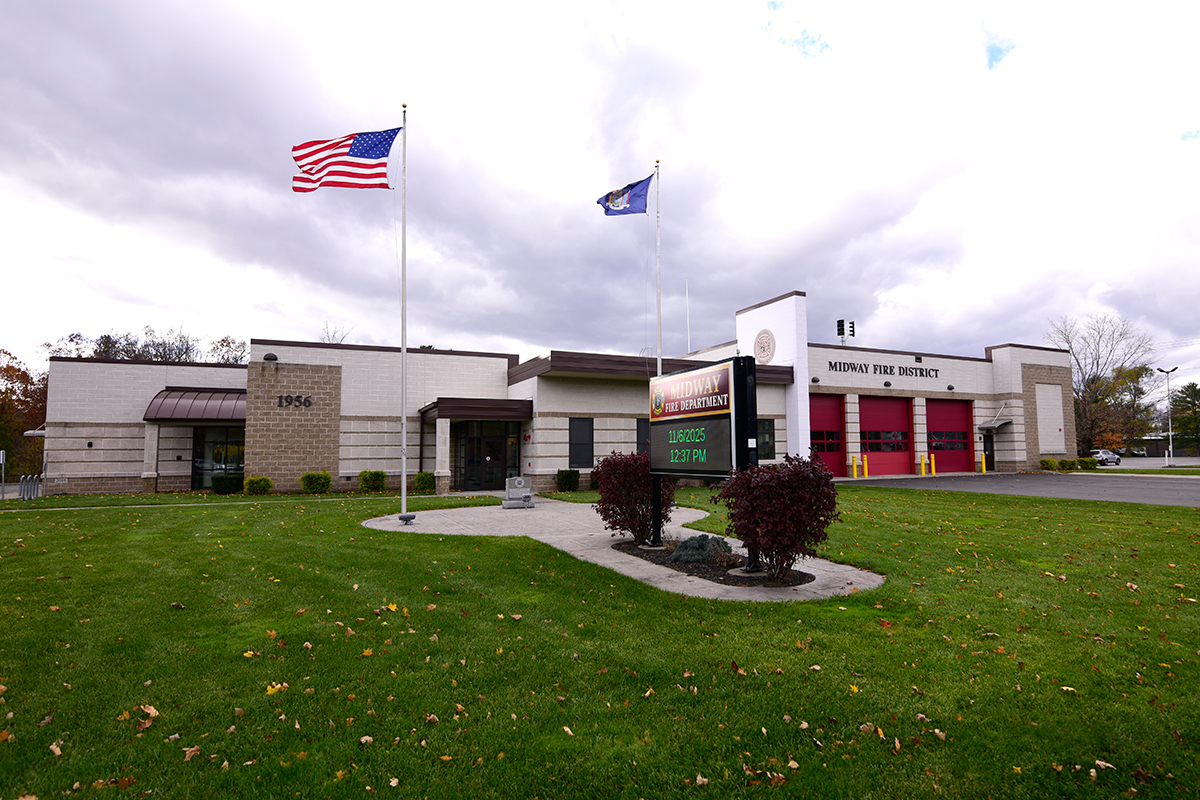 A fire station with three red garage doors, two flagpoles flying the U.S. and state flags, a sign with electronic display, and a well-kept lawn under a cloudy sky.