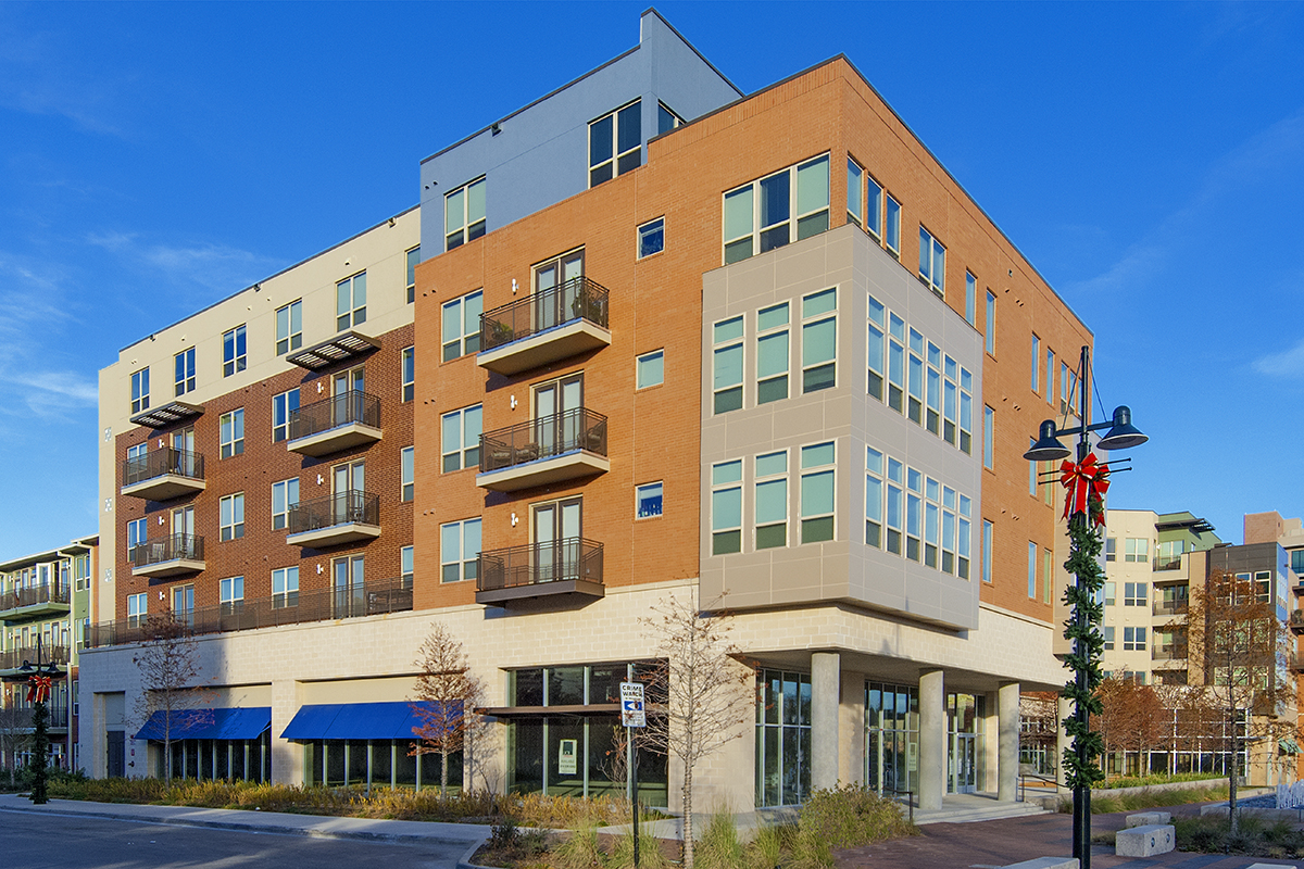 A modern, four-story apartment building with brick and light-colored siding, balconies, large windows, and ground-floor retail space under a clear blue sky. A streetlamp is decorated with a holiday wreath and ribbon.
