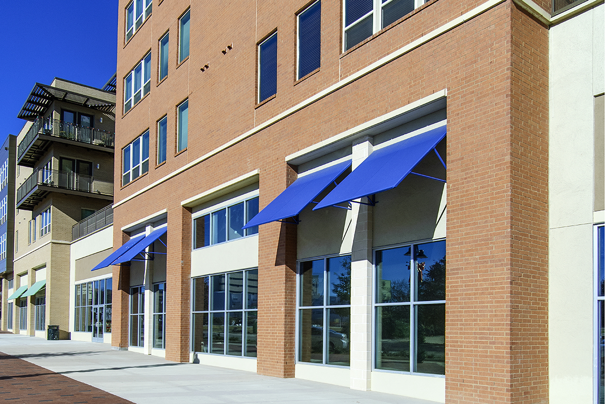 A brick commercial building with large glass windows and blue awnings. Shadows from the awnings fall on the sidewalk. Another building with balconies is visible to the left. The sky is clear and blue.